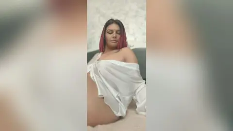 Elegant woman with pink hair in a white silk shirt relaxing in a bright, dreamy bedroom setting.