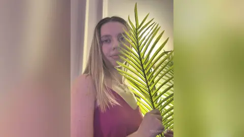 Young woman in a pink tank top posing with a green palm leaf in a bright, warm indoor setting.