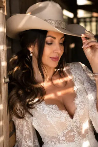 Beautiful woman in white lace wedding dress and beige cowboy hat posing in warm sunlight.
