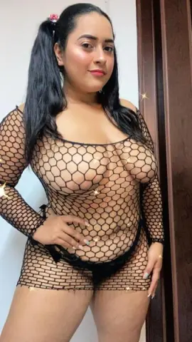 Curvy woman in a black fishnet mini dress posing indoors with a glamorous aesthetic.