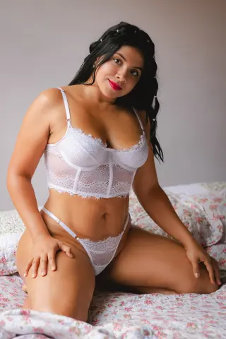 Curvy woman in white lace lingerie sitting on a floral bed in a bright, cozy bedroom setting.