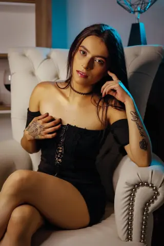 Young woman in a black lace off-the-shoulder dress sitting on a white armchair in moody lighting.