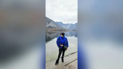 Woman in a cozy blue faux fur coat and black leggings standing by a scenic mountain lake under a cloudy sky.
