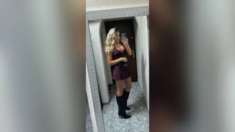 Blonde woman in a burgundy mini dress and black boots taking a mirror selfie in a hallway.