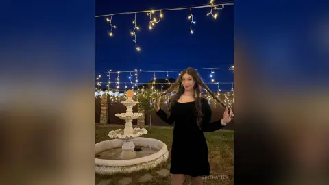 Young woman in a black dress holding her long hair by a tiered fountain under glowing string lights at night.