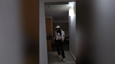 Young woman in a white crop top and black pants taking a mirror selfie in a dimly lit bedroom.