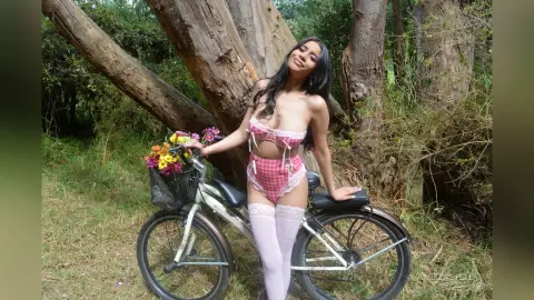 AmeliaStell poses naked or in light pink checkered lingerie while sitting on a bicycle in a wooded outdoor setting.