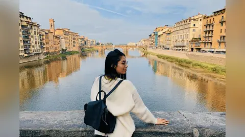 LucyMartin leans against a stone wall overlooking a river, wearing a white sweater and a black backpack.