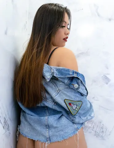 Young woman with long brown hair wearing a denim jacket slung off her shoulders against a white textured wall.