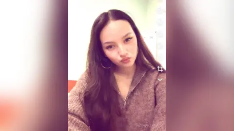 Young woman with long brown hair wearing a cozy brown knit sweater in a bright indoor setting.