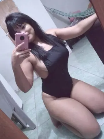 Curvy woman in a black bodysuit taking a casual mirror selfie in a bright room.