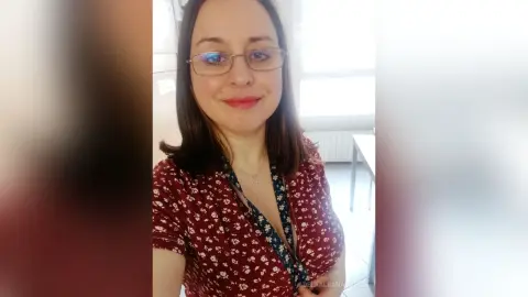 AdelaAlbaNatural — Smiling woman with glasses wearing a floral print blouse in a bright indoor setting.