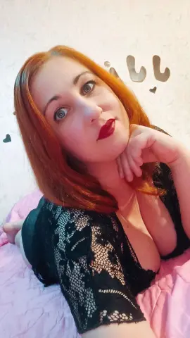 Jill_Stevens poses with her hand under her chin, wearing a black lace top that shows off her cleavage and red hair.