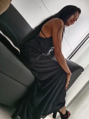 Elegant woman in a long black evening gown sitting on a grey sofa in a bright modern room.