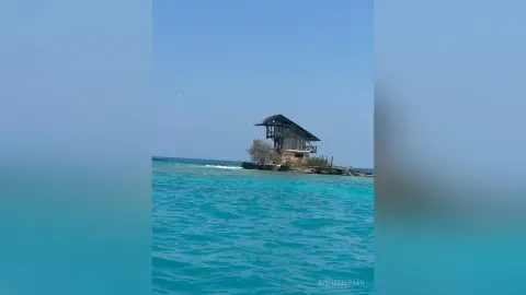 A rustic wooden beach hut sits on a small rocky island in the middle of calm turquoise ocean waters under a clear sky.