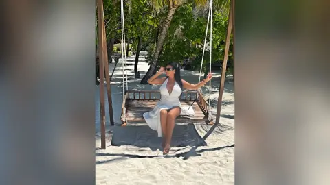 MissyJolie sits on a wooden swing in a white sundress, posing outdoors near palm trees on a sandy beach.