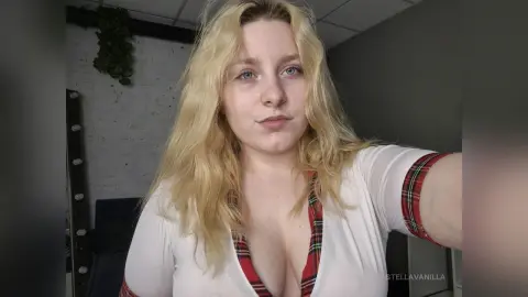 StellaVanilla with long blonde hair wearing a white shirt with red plaid trim, showing cleavage in a close-up selfie.