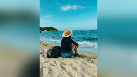 Woman in a black shirt and denim shorts sitting on a sandy beach facing the blue ocean under a clear sky.