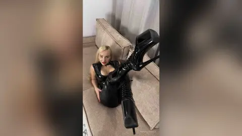 Blonde woman in a black latex bodysuit and high heel platform boots lounging on a beige sofa.