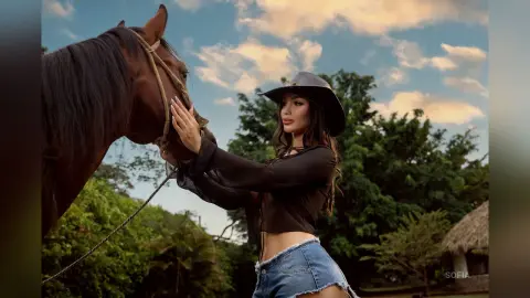 Young woman in a black top and cowboy hat petting a brown horse in a lush outdoor garden setting.