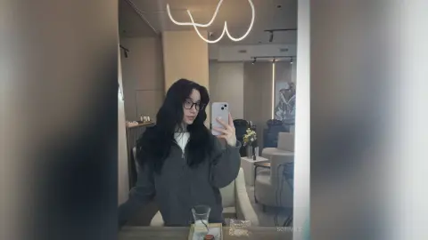 SofiMils poses for a mirror selfie wearing a grey zip-up sweater and black glasses, showing off her long, dark wavy hair.