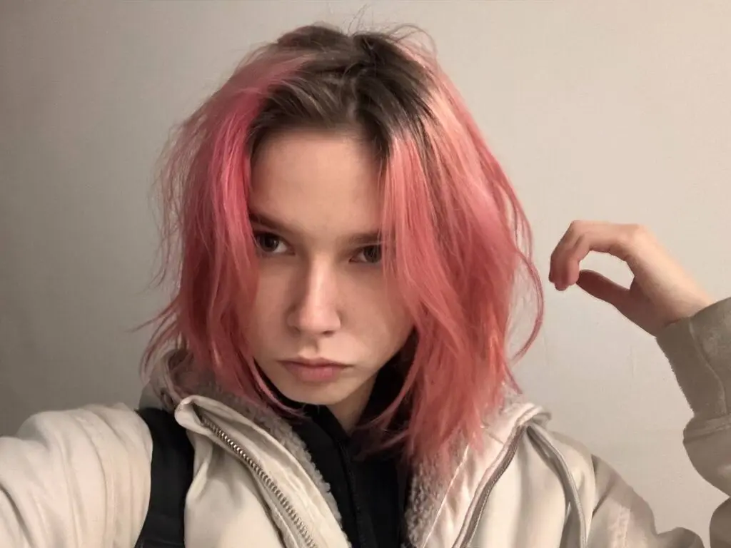 Young woman with pink hair wearing a cream fleece jacket in a minimalist indoor setting.