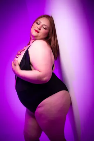 Curvy woman in a black bodysuit posing confidently against a vibrant purple studio background.