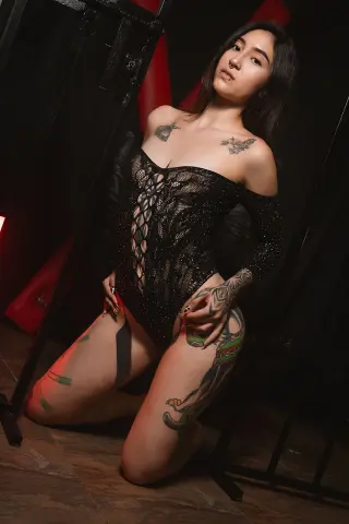 MeganPressley poses in a black lace bodysuit, showing off her tattoos and long dark hair in a dimly lit room.