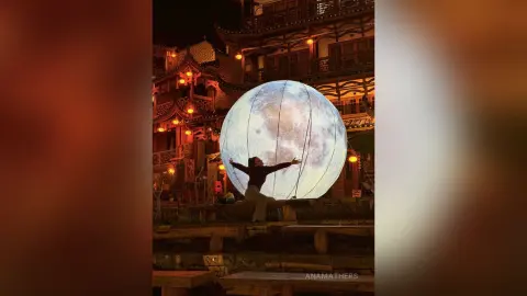 Woman in casual attire spreading arms toward a giant glowing moon lantern in a traditional Asian village at night.
