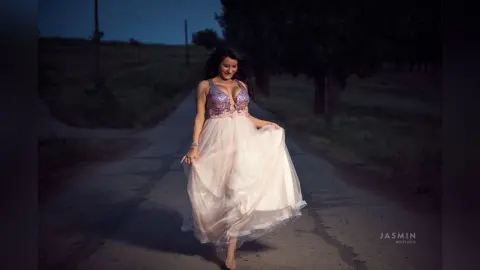 MissyJolie stands on a dark path wearing a purple sequined bustier top and a long, flowing light pink tulle skirt.
