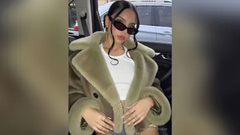 Stylish woman in a fuzzy olive faux fur coat and white tank top sitting in a car wearing sunglasses.
