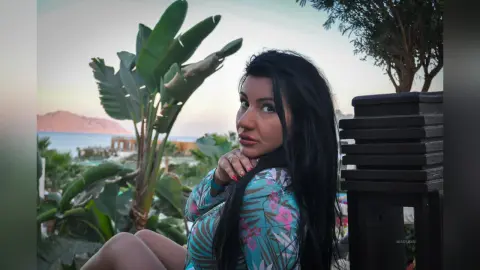 MissyJolie poses outdoors with long black hair, wearing a blue floral print top or dress near tropical green plants.