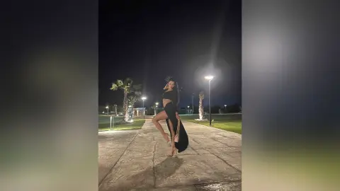 NicoleBella poses outdoors at night in a black crop top and long skirt with a high slit, standing on a concrete path near palm trees.