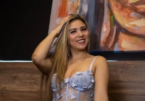 Smiling blonde woman in a light blue floral lace bodysuit posing in a cozy indoor setting.