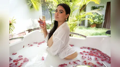 NatalyBloom sits in a white bathtub filled with milk and rose petals, wearing an open white shirt and holding a glass of wine.