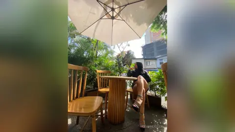 Young woman in a black outfit sitting at an outdoor cafe table under a white patio umbrella on a sunny day.