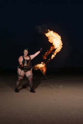 Curvy performer in black bodysuit performing fire breathing stunt under a dark night sky.