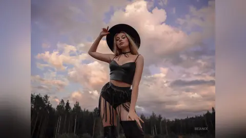BeaNix poses in a black tank top and fringed skirt with a black hat against a cloudy sky.