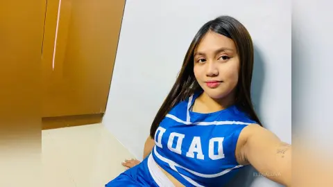 Young woman in a blue and white sleeveless crop top smiling indoors against a plain wall.
