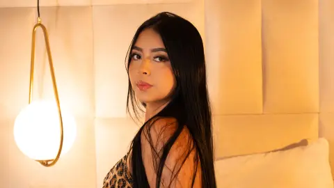 Young woman with long dark hair in a leopard print top posing in warm, ambient bedroom lighting.