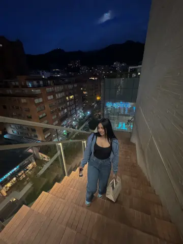 IndiraJhons stands on a set of outdoor stairs wearing a black tank top and blue jeans, holding a small bottle and a white bag.
