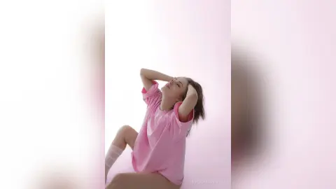 NoellaGlowinski wearing a pink t-shirt, running her hands through her hair while sitting against a white background.