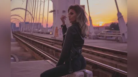 AnaMathers poses in a black leather jacket and black pants while sitting on a ledge next to train tracks at sunset.