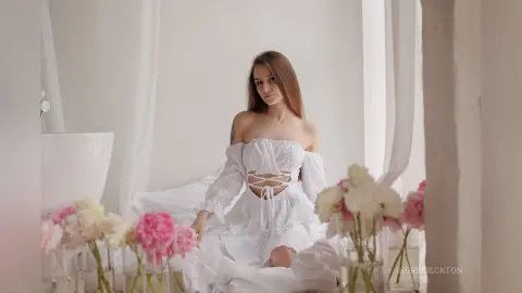 IngridBeckton poses in a white lace off-the-shoulder top and skirt, sitting on white bedding surrounded by pink and white flowers.