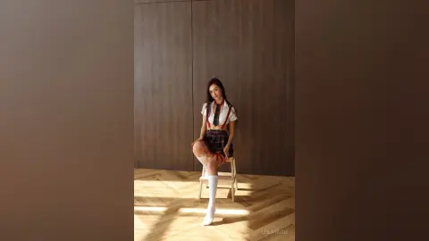 Young woman in a white blouse and plaid skirt sitting on a chair in a sunlit minimalist room.