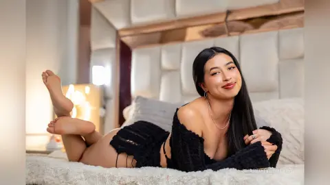 Smiling young woman in a black knit outfit relaxing on a cozy white bed in a warm, bright bedroom setting.