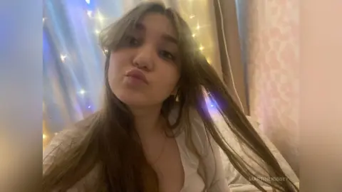 MartineKibbey pouts her lips while letting her long brown hair fall over her shoulders in a room with soft string lights.
