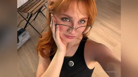 AlbertaCorrell is a redhead wearing glasses and a black tank top, posing for a selfie in a room with wood floors.