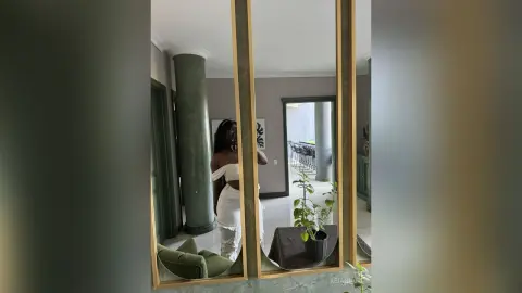 KeianHoz stands in a room wearing a white off-the-shoulder top and white pants, reflected in a gold-framed mirror.