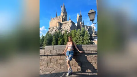 Young woman in a white crop top and denim shorts posing by a stone wall at the magical Hogwarts castle.